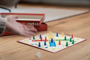 Main lançant un dé à côté d'un plateau de jeu de parcours en croix, avec pions coniques rouges, bleus, jaunes et verts, sur une table en bois