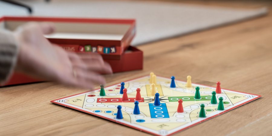 Main lançant un dé à côté d'un plateau de jeu de parcours en croix, avec pions coniques rouges, bleus, jaunes et verts, sur une table en bois